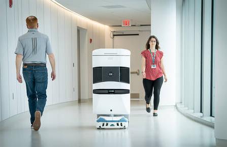 A TUG robot is seen traveling a hospital hallway with two people walking on either side of the robot.